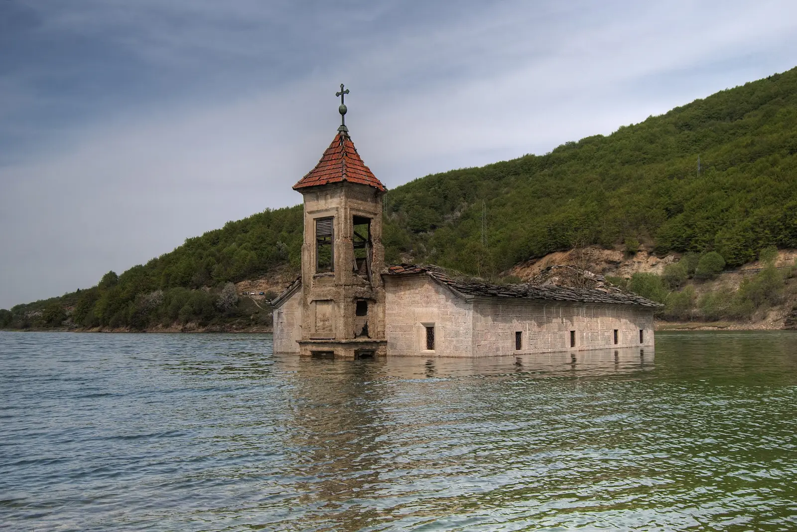Потопената црква „Свети Никола“ во Мавровското Езеро, со камбанаријата над водата и зеленото ридо позади